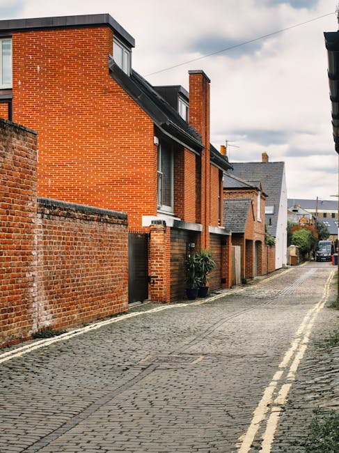 A row of brick terraced houses with chimneys and small front gardens, situated on a narrow street under a clear blue sky. In the foreground, there is a parking area with several vehicles, including a 