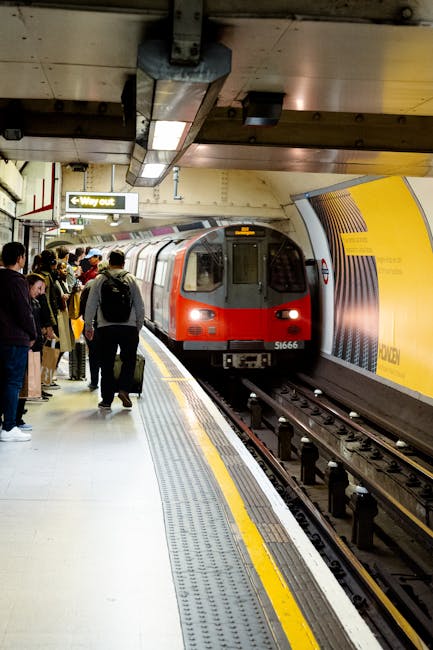 In a busy underground train station, a red and grey train with the number 51666 approaches the platform where passengers, including men and women, are waiting to board. Some passengers are standing near the edge of the platform, holding luggage or backpacks, while others are engaged in conversation or checking their phones. The platform has tactile paving strips along the edge in yellow and grey, providing guidance for visually impaired travelers. Overhead, signs indicate directions and exits, with one sign showing 'Way out'. The ceiling is industrial with visible metal beams, lighting fixtures, and ventilation. The environment appears well-lit and organized to facilitate home relocation or furniture transport logistics typical of station transfers associated with house removals. Man With a Van Redbridge's removals services may coordinate such transit steps during moving processes, ensuring safe and efficient transport of belongings from station to destination.