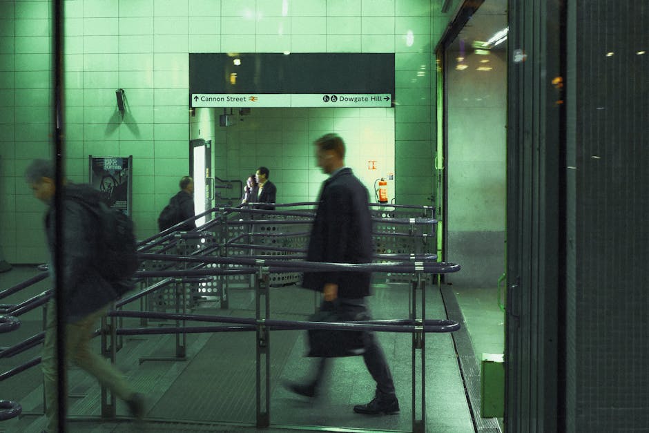 Inside a London underground station, green-tiled walls surround a spacious area with metal railings guiding passengers through a designated walkway. Several commuters are visible, including two men wearing dark business suits who are walking in opposite directions; one man is carrying a black briefcase, while another is seen with a backpack. The lighting is artificial, casting a neutral tone across the platform. In the background, a sign indicates directions towards Cannon Street and Dowgate Hill, with the station's name partially obscured. The scene depicts a busy moment during a typical day of mobility and travel, which relates to the broader context of home relocation and moving services, such as those offered by Man With a Van Redbridge, including furniture transport, packing, and loading process in urban transportation environments.