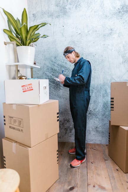 A man dressed in a dark blue jumpsuit and red slippers is standing inside a room with a light textured wall and wooden flooring, surrounded by several cardboard boxes used for packing and moving household items. He is holding a blue pen and appears to be taking notes or checking a clipboard, indicating a furniture transport or home relocation process. To his left, there is a large white plant pot with a green leafy plant placed on a white shelf attached to the wall. The boxes, some with labels and others sealed, are stacked and positioned near him, suggesting the packing and moving phase typical of house clearance or relocation services. The scene is well-lit, emphasizing the practical environment associated with moving logistics. This image relates to the services offered by Man With a Van Redbridge, highlighting the careful handling and organization involved in furniture transport and house removals.