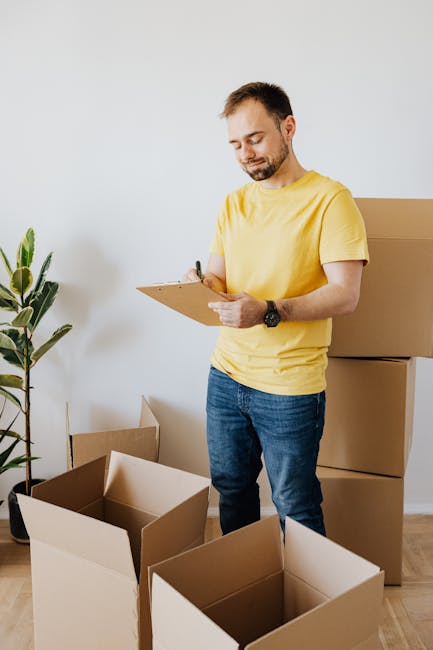 A man dressed in a dark blue jumpsuit and red slippers is standing inside a room with a light textured wall and wooden flooring, surrounded by several cardboard boxes used for packing and moving household items. He is holding a blue pen and appears to be taking notes or checking a clipboard, indicating a furniture transport or home relocation process. To his left, there is a large white plant pot with a green leafy plant placed on a white shelf attached to the wall. The boxes, some with labels and others sealed, are stacked and positioned near him, suggesting the packing and moving phase typical of house clearance or relocation services. The scene is well-lit, emphasizing the practical environment associated with moving logistics. This image relates to the services offered by Man With a Van Redbridge, highlighting the careful handling and organization involved in furniture transport and house removals.