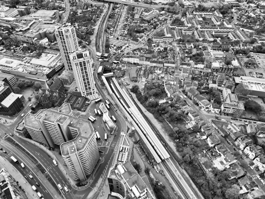 A black and white aerial photograph of a densely built urban area showing multiple high-rise office buildings and smaller residential houses. Prominent in the image is a railway station with train platforms and tracks, partially visible near the center, suggesting a busy transportation hub. Adjacent to the station, a variety of vehicles are parked or moving along the roads, with some buses visible at a bus stop close to the station. The surrounding roads curve through the cityscape, with traffic flowing in different directions. Green spaces and trees are dispersed among the buildings, providing contrast with the concrete structures. The lighting indicates daytime with even illumination across the scene. This image captures a snapshot of the city's infrastructure, typical of a large metropolitan area, suitable for illustrating urban development, transport systems, or home relocation logistics. Man With a Van Redbridge occasionally references moving and packing processes in similar contexts, although no specific moving activity is visible in this aerial shot.