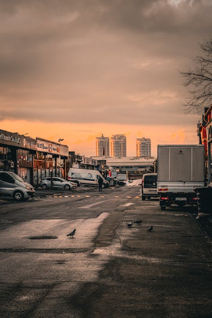 A view of a commercial parking area during sunset with a cloudy sky, featuring several parked vehicles including a white van and a small car. The parking lot is surrounded by low-rise buildings with shopfronts, some with signage, and a few pedestrians are visible. In the background, three taller modern high-rise buildings are seen against the orange and pink hues of the evening sky. The surface of the parking area appears wet, with puddles reflecting the surroundings. This scene illustrates urban logistics and transportation settings that could relate to house removals and furniture transport, complementing services offered by Man With a Van Redbridge in Ilford IG1 for street-by-street relocations.