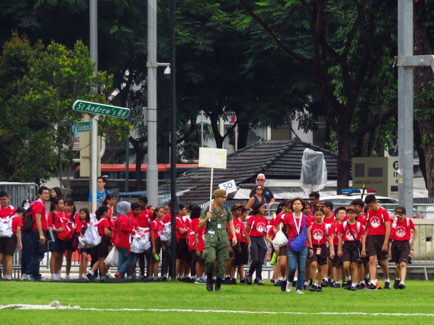 A group of schoolchildren dressed in red t-shirts and dark shorts, carrying backpacks and white plastic bags, are walking in a line across a grassy area, guided by a uniformed personnel holding a sign with '50' on it. They are outdoors, near a street corner with trees, street signs, and buildings in the background, including a small structure with a tiled roof. The scene is well-lit with natural daylight, and the children appear to be participating in an organized event or school outing. This setting reflects a typical environment for home relocation or community-based activities, with the focus on group movement and transportation logistics, as part of a moving or packing process, which manwithavanredbridge.co.uk’s removals service might support.