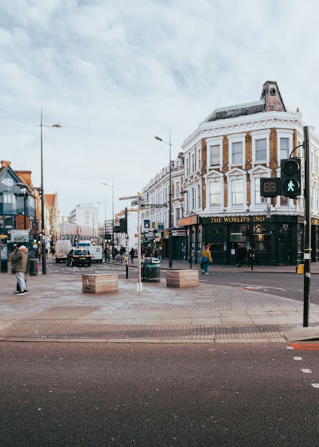 A busy urban street scene on Ilford High Road featuring a corner building with a white facade, large windows, and a sign reading 'The World's End' at ground level. In the foreground, a pedestrian crossing with a green walking signal is visible, along with several people walking on the pavement. On the left, there are shops and storefronts, while on the right, a road with moving vehicles can be seen. The sky is partly cloudy with patches of blue. The scene captures typical city life, relevant to house removals and packing services offered by Man With a Van Redbridge for home relocation purposes.