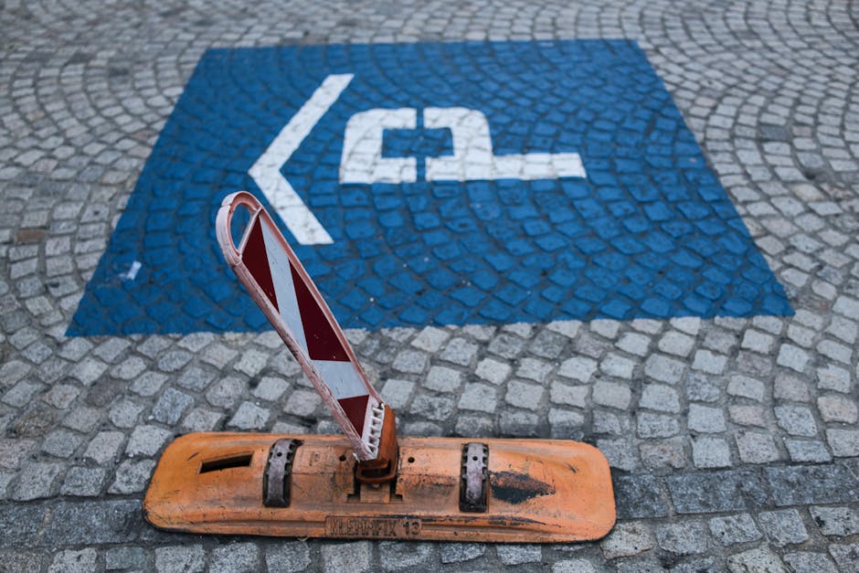 A pavement area in a residential or urban setting features a blue painted parking space designated for disabled parking, marked with a white wheelchair symbol and an arrow pointing left. In front of the space, a traffic barrier with red and white reflective stripes is mounted on an orange base, which appears slightly worn and dirty. The barrier is resting against the base, tilted at an angle. The surrounding ground consists of cobblestone paving, and the scene is illuminated with natural daylight, indicating an outdoor environment. The image relates to house removals and moving logistics, demonstrating the importance of compliance with parking restrictions and clearance during home relocation or furniture transport, with [COMPANY_NAME] involved in managing such logistics discreetly.