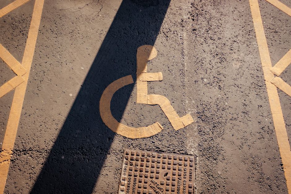 A pavement area in a residential or urban setting features a blue painted parking space designated for disabled parking, marked with a white wheelchair symbol and an arrow pointing left. In front of the space, a traffic barrier with red and white reflective stripes is mounted on an orange base, which appears slightly worn and dirty. The barrier is resting against the base, tilted at an angle. The surrounding ground consists of cobblestone paving, and the scene is illuminated with natural daylight, indicating an outdoor environment. The image relates to house removals and moving logistics, demonstrating the importance of compliance with parking restrictions and clearance during home relocation or furniture transport, with [COMPANY_NAME] involved in managing such logistics discreetly.