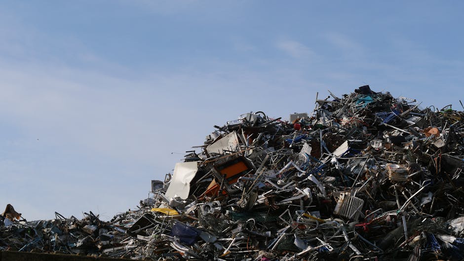 A large pile of assorted scrap metal, broken appliances, and discarded household items, including metal frames, twisted wires, and mangled furniture, stacked outdoors against a clear blue sky. The debris consists of damaged metal objects and plastic parts, with some items partially covered in fabric or plastic wrapping. The pile extends upward and appears to be an accumulation of waste material awaiting recycling or disposal, typical of waste management or recycling sites. This scene illustrates the final stages of house removal or clearance processes, where construction debris, unwanted appliances, and other bulky waste materials are gathered. Man With a Van Redbridge is likely involved in transporting or managing such waste as part of their comprehensive removals and relocation services, ensuring proper handling of household items and bulk waste within home relocation projects or bulky waste management in Redbridge.