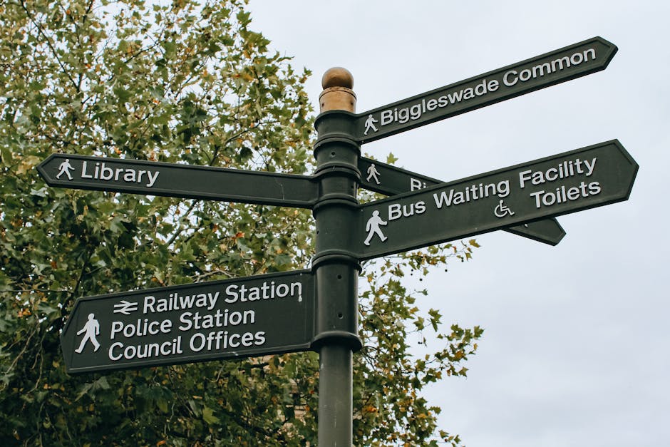 A black multi-directional signpost situated outdoors in a location with green foliage and an overcast sky background. The signpost features multiple rectangular signs with white text and icons, indicating directions to local amenities such as Biggleswade Common, Library, Railway Station, Police Station, Council Offices, Bus Waiting Facility, and Toilets. The signs are mounted on a central pole with a rounded top, and some signs have walking figures or wheelchair icons to denote pedestrian routes and accessible facilities. The image captures a scene relevant to community navigation and urban signage, which may be useful in the context of house removals, moving logistics, or local area orientation, as referenced by the topic 'Redbridge council removal permits.' Man With a Van Redbridge may encounter or utilize such signage during home relocation or furniture transport processes, emphasizing the importance of understanding local signage for efficient moving services.
