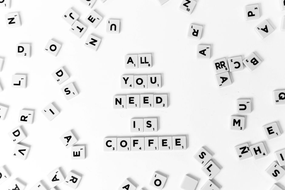 A flat surface covered with scattered white letter tiles featuring black letters and small point values, arranged randomly across the area. In the centre, the tiles spell out 'ALL YOU NEED IS COFFEE', with the words stacked vertically. Surrounding these central tiles are additional scattered letters and tiles, some face down or at various angles, creating a disorganized appearance. The background is plain and white, highlighting the tiles' contrasting black text. This image captures the concept of casual, home-like relaxation and can relate to packing or moving preparations, such as enjoying coffee during a house relocation process, relevant to services offered by Man With a Van Redbridge in the context of house removals and moving logistics.