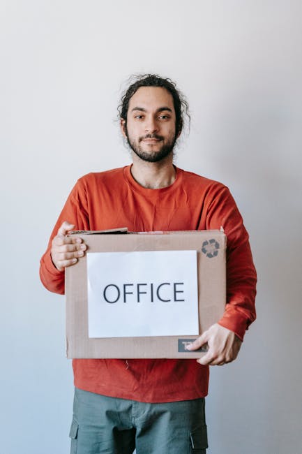 A man and a woman standing indoors against a plain white wall, with the man holding a cardboard box labeled 'OFFICE' wrapped in clear packing tape. The man has curly dark hair, a beard, and is dressed
