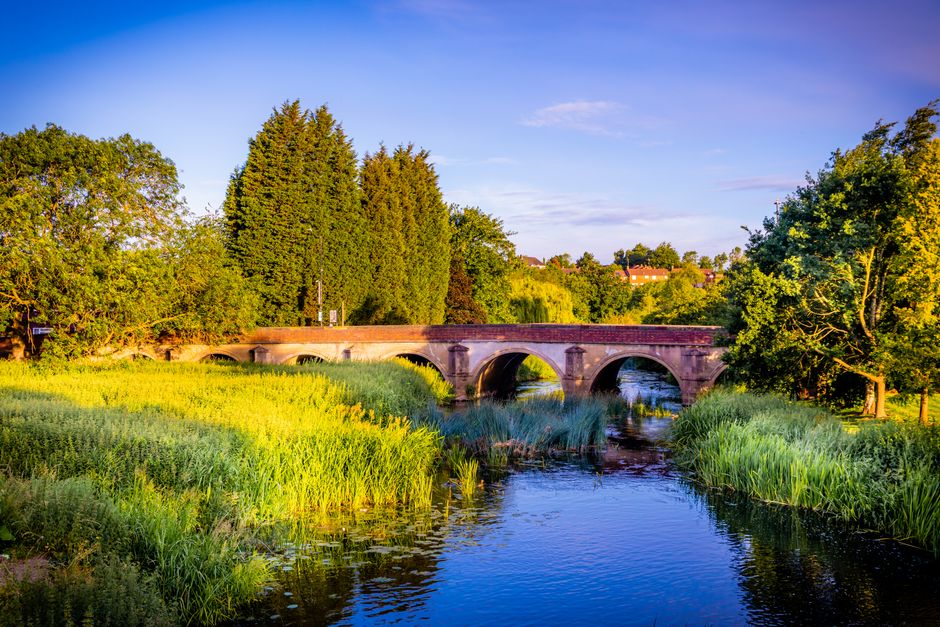 A stone arch bridge spans a narrow river or stream in a scenic outdoor setting, with lush green vegetation and tall trees on both sides. The bridge has multiple rounded arches supporting a flat roadway, and the scene is illuminated by natural sunlight, suggesting late afternoon or early evening. The water below reflects the sky and surrounding foliage, adding to the tranquil atmosphere. In the background, there are additional trees and distant buildings, with a clear blue sky overhead. The image captures the peacefulness of the landscape and the natural environment, with the bridge serving as a structural element amid the greenery. This setting is suitable for illustrating home relocation or moving logistics, with the focus on transportation and scenic routes often associated with property moves, such as those managed by Man With a Van Redbridge or other UK removals services.