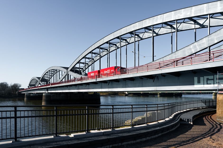 A large, modern steel and glass bridge spanning a body of water under a clear blue sky. The bridge features multiple curved arches and a red railing along the edges. A red vehicle, possibly a van or moving truck, is visible on the bridge, aligned with the concept of furniture transport or home relocation. Below the bridge, there is a viewing walkway with black metal railings and curved paving, suggesting a scenic area for pedestrians. The quiet water reflects some of the bridge's structure, and nearby trees are visible in the background, indicating an outdoor setting. This scene highlights the infrastructure involved in relocation logistics, with a focus on moving vehicles travelling across key transport routes, supporting services like those offered by Man With a Van Redbridge for house removals and furniture transport.