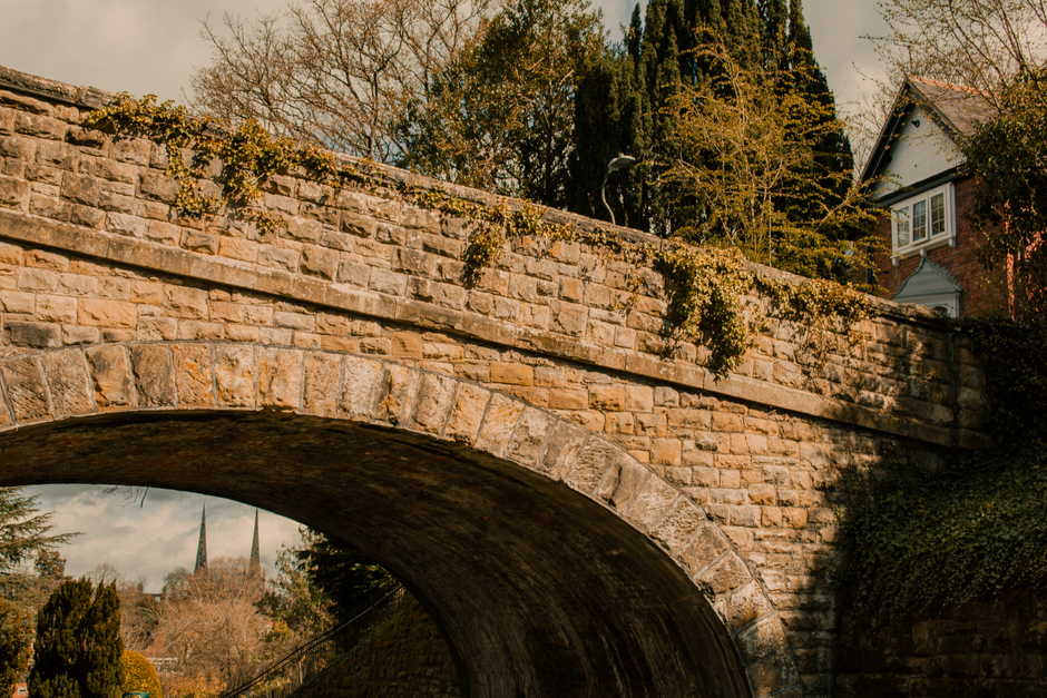 A stone arch bridge spans a narrow river or stream in a scenic outdoor setting, with lush green vegetation and tall trees on both sides. The bridge has multiple rounded arches supporting a flat roadway, and the scene is illuminated by natural sunlight, suggesting late afternoon or early evening. The water below reflects the sky and surrounding foliage, adding to the tranquil atmosphere. In the background, there are additional trees and distant buildings, with a clear blue sky overhead. The image captures the peacefulness of the landscape and the natural environment, with the bridge serving as a structural element amid the greenery. This setting is suitable for illustrating home relocation or moving logistics, with the focus on transportation and scenic routes often associated with property moves, such as those managed by Man With a Van Redbridge or other UK removals services.