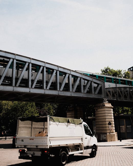 A white flatbed van with a partially covered cargo area is parked on a paved area beneath a railway bridge. The van's rear doors are open, revealing a load of cardboard boxes and packing materials inside the cargo space. A green fabric cover is draped over part of the load to secure the items. The bridge overhead is constructed of metal trusses supported by stone pillars, with a train visible on the elevated track above. The surrounding environment includes some trees and urban features, indicating a city or town setting. This scene depicts a typical loading process during home relocation or furniture transport, with the van positioned for ease of loading or unloading. The presence of packaging materials suggests careful packing and moving activities, consistent with professional removals services by Man With a Van Redbridge, as part of a same-day removal operation covered on the webpage about Redbridge house removals.