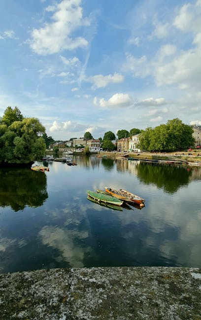 A scenic view of a calm river reflecting the partly cloudy sky above, with several small boats moored along the bank. On the far side of the river, there are rows of residential buildings and lush green trees lining the waterfront. The image captures the peaceful environment typical of a house relocation area, where moving companies like Man With a Van Redbridge may operate. The presence of boats and the natural surroundings suggest an accessible, tranquil setting suitable for local removals and home moving logistics. The overall scene is well-lit with natural daylight, emphasizing the serene atmosphere of the river and its nearby housing district, which may be part of a residential move or local furniture transport process.