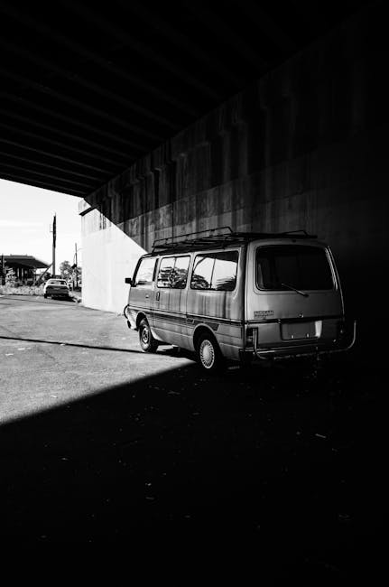 A black and white photograph of a small van parked underneath a large concrete overpass. The van is positioned on a paved surface with a clear area surrounding it, partly shaded by the shadow of the overpass structure. The van appears to be a typical vehicle used for home relocation or furniture transport, with a boxy shape and windows along the sides. The background shows a few distant vehicles and structures, indicating an industrial or urban setting. This image depicts the initial stage of a moving process, where a vehicle, associated with [COMPANY_NAME], is ready for loading or unloading furniture and packing supplies in preparation for a house removal, highlighting the logistics involved in packing, loading, and transporting household items within a professional moving service.