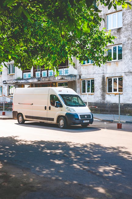 A white moving van with a high roof and black bumper is parked on a street adjacent to a residential building with multiple windows. The van's side doors are closed, and it is positioned near the pavement, partially shaded by large green tree foliage overhead. The building behind the van appears weathered, with visible signs of age on its exterior walls, and features balconies with decorative railings, some of which display snowflake decorations. The scene suggests a home relocation or furniture transport process, with the van ready for loading or unloading items. The surrounding area is calm with minimal street traffic, and the environment indicates a typical urban residential setting, supporting professional removals services like those offered by Man With a Van Redbridge.