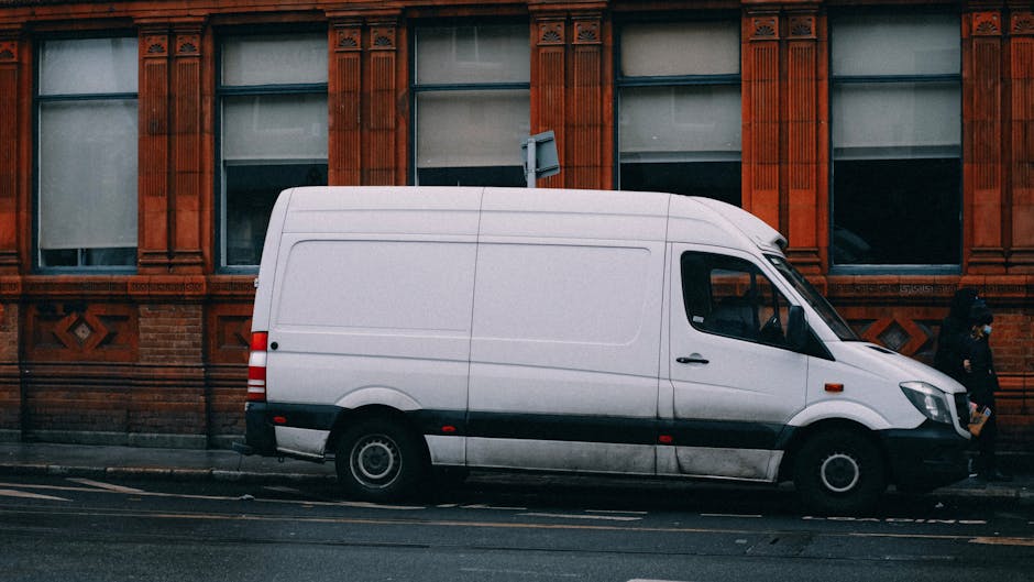 A white commercial van with black side stripe is parked along the pavement outside a red-brick building with decorative window surrounds. The van is positioned parallel to the curb on an urban street, with its sliding side door closed. The vehicle is surrounded by a loading area suitable for home relocation activities, and the street has visible road markings. In the background, a person dressed in dark clothing is seen walking past, and another individual is partially visible near the rear. The scene suggests a furniture transport or moving process, with the van ready for loading or unloading belongings during packing and moving activities. Man With a Van Redbridge, a removals service, may be involved in coordinating this logistics, supporting efficient property moves or house relocations in the area.