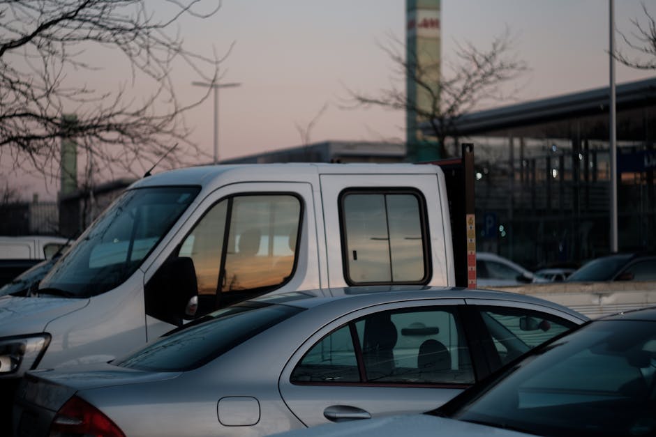 A white commercial van, partially visible, parked in a lot during dusk with already loaded items, visible through its open rear doors, including cardboard boxes and packing materials, which are being prepared for a home relocation. In front of the van, a silver sedan is parked, with its windows reflecting the soft evening light. The background shows other vehicles, some trees with bare branches, and a modern building with large glass windows and a canopy roof. The scene depicts the loading process typical of furniture transport and packing during house removals, supported by Man With a Van Redbridge, in an environment with natural lighting and clear visibility of loading equipment and objects.