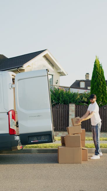 A man wearing a white t-shirt and jeans is standing next to an open white cargo van on a suburban street during daylight. He is in the process of loading or unloading medium-sized cardboard boxes, wit